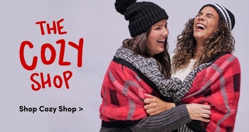 Two women are laughing and embracing each other while wrapped in a red plaid blanket They are dressed warmly wearing knit hats and cozy sweaters creating a cheerful and friendly winter atmosphere The text on the left side reads The Cozy Shop with a prompt below saying Shop Cozy Shop The image conveys a warm and inviting feel encouraging holiday shopping for comfortable winter wear