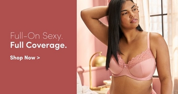 Confident woman wearing a pink lace-trimmed bra standing in a softly lit bedroom with a pink wall and gold decor. Text reads Full-On Sexy. Full Coverage. Shop Now.