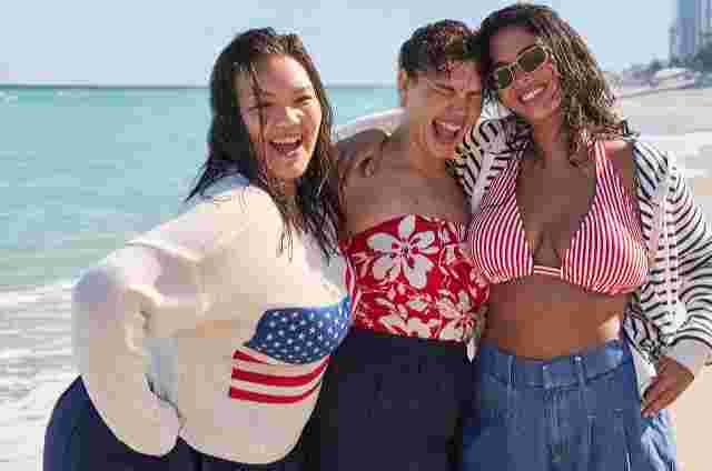 Three smiling women standing arm-in-arm on a sunny beach. One wears a white sweater with an American flag, one wears a red floral swimsuit, and one wears a red striped bikini top with a hoodie and denim shorts. Ocean waves and city skyline in the background