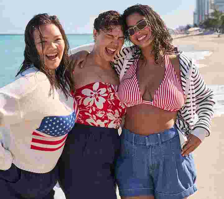 Three smiling women standing arm-in-arm on a sunny beach. One wears a white sweater with an American flag, one wears a red floral swimsuit, and one wears a red striped bikini top with a hoodie and denim shorts. Ocean waves and city skyline in the background
