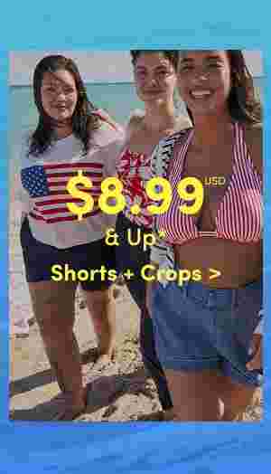 Three smiling women walking on the beach wearing summer outfits including a flag sweater, floral swimsuit, and red striped bikini top with denim shorts. Overlaid text reads 8.99 USD and up, Shorts + Crops