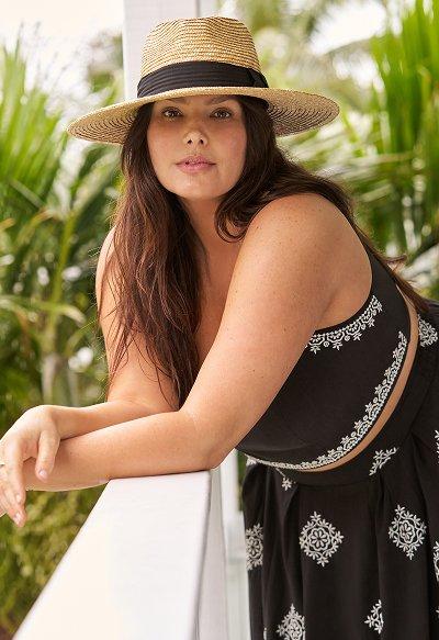 Woman leaning on a white railing wearing a wide-brimmed straw hat and a black sleeveless dress with white embroidered details. Tropical background with palm leaves.