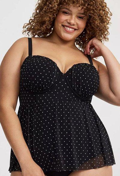 Smiling woman wearing a black polka-dot swim top with a flowy, sheer overlay and adjustable straps, posing in front of a light background.