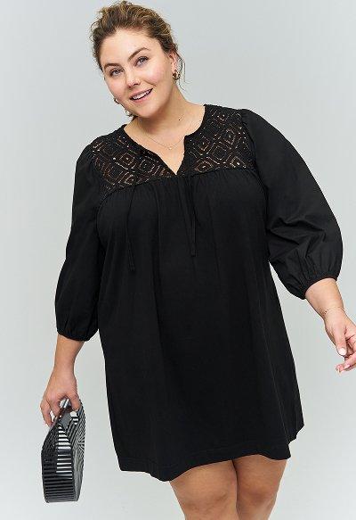 Woman smiling and holding a striped handbag, wearing a black long-sleeve mini dress with puff sleeves and lace detailing at the neckline.