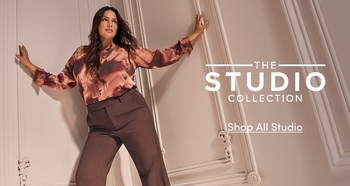 A model wearing a patterned satin blouse and dark trousers poses against a white paneled wall. Text on the right reads “The Studio Collection” with a call to action “Shop All Studio.”