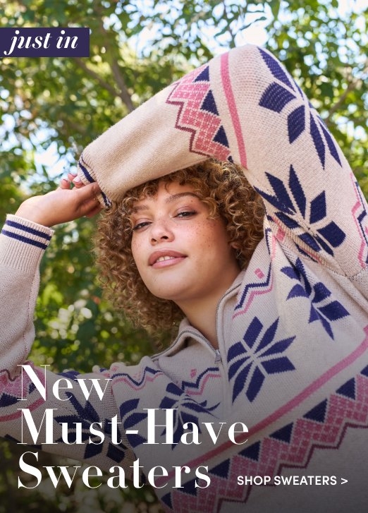'Woman with curly hair poses outdoors in front of green trees wearing a light beige sweater with a quarter zip and a navy and pink geometric snowflake pattern. White text at the top left reads just in. Large white text at the bottom left reads New Must-Have Sweaters. Smaller white text at the bottom right reads SHOP SWEATERS >.'
