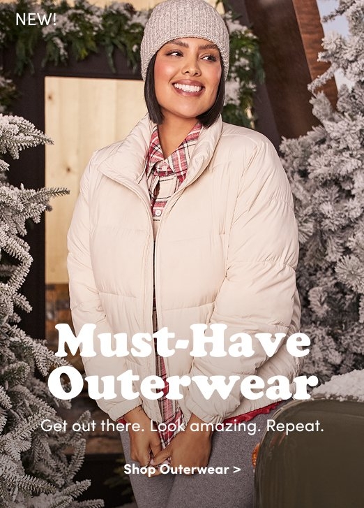 'Model with short dark hair smiling while standing between snow-frosted trees, wearing a beige knit beanie, cream puffer jacket, and red plaid shirt. White text reads New! Must-Have Outerwear. Below it: Get out there. Look amazing. Repeat. A white button reads Shop Outerwear >.'