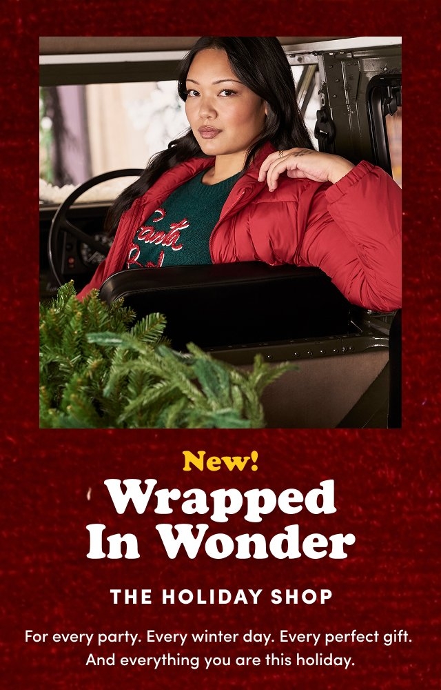 Woman sitting in the driver seat of a vehicle wearing a red puffer jacket over a green sweater with red and white text that reads 'Santa Baby', resting her arm on the seat back with greenery branches in the foreground and a blurred snowy background outside the window. Below, on a dark red textured background, centered yellow and white text reads 'New! Wrapped In Wonder THE HOLIDAY SHOP For every party. Every winter day. Every perfect gift. And everything you are this holiday.'