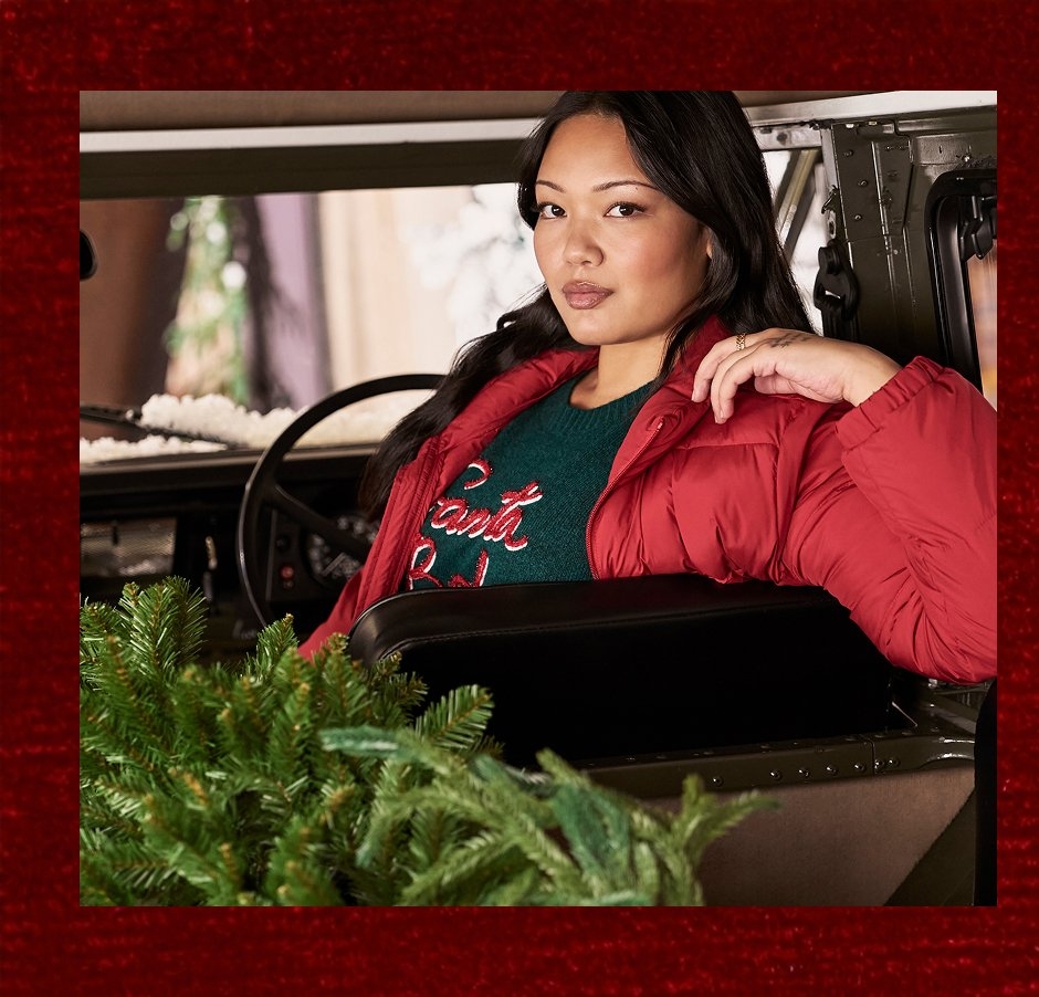 Woman sitting in the driver seat of a vehicle wearing a red puffer jacket over a green sweater with red and white text that reads 'Santa Baby', resting her arm on the seat back with greenery branches in the foreground and a blurred snowy background outside the window.