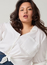 A close-up of a woman with long dark wavy hair, looking toward the camera with her lips slightly parted. She wears small gold hoop earrings and an oversized white button-down shirt with a chest pocket and long sleeves rolled at the cuffs, tied at the hem, over dark wash denim jeans visible at the bottom. The background is a plain light gray.