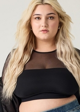 A close-up of a woman with long wavy blonde hair looking directly at the camera. She wears small stud earrings and a black ribbed cropped bandeau top layered under a sheer black mesh long-sleeve top, with a black faux leather jacket visible at the shoulders and dark wash denim jeans at the bottom. The background is a plain light gray.