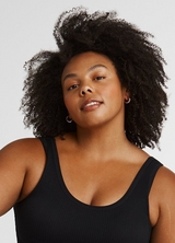 A close-up of a woman with voluminous natural dark curly hair, looking toward the camera with a soft expression. She wears small gold hoop earrings and a black ribbed scoop-neck tank top. The background is a plain light gray.