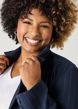 A close-up of a woman with short curly dark hair with caramel highlights, smiling widely. She wears small gold hoop earrings, a silver ring, a white V-neck top, and a navy blue pinstriped blazer, which she holds open at the collar with one hand. The background is a plain light gray.