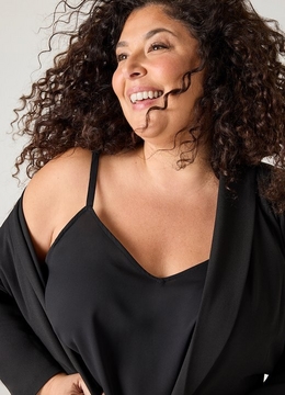 A woman with long voluminous curly dark brown hair laughs with her head tilted back and to the side.A woman with long voluminous curly dark brown hair laughs with her head tilted back and to the side. She wears a delicate gold chain necklace, a black satin V-neck cami with thin spaghetti straps, and a black wide-sleeve open cardigan or wrap draped off her shoulders. The background is a plain light gray.