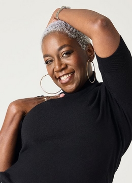 A woman with short natural gray and white hair smiles broadly with one arm raised behind her head. She wears large gold hoop earrings, a bracelet, a ring, and a black cold-shoulder knit top. The background is a plain light gray.