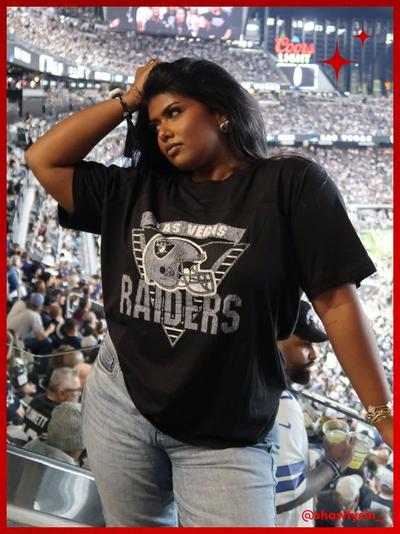 'a woman with long dark hair poses in a crowded football stadium wearing a loose black las vegas raiders graphic t-shirt with a large silver helmet logo across the chest paired with light-wash jeans with her left hand resting on her head and her right arm bent at her side red star graphics appear in the top right corner and her username @shawtysin is printed in red in the bottom right corner'