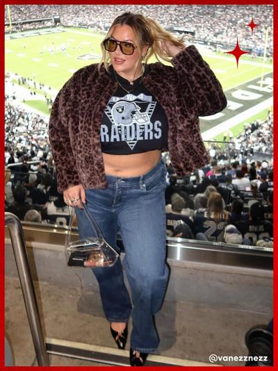 'A woman stands in a stadium suite with a crowded football field behind her, wearing oversized square sunglasses, a cropped black Las Vegas Raiders graphic tee, mid-wash flare jeans, black pointed-toe heels with gold hardware, and a cropped leopard-print faux fur jacket; she holds a clear boxy handbag with a chain strap in her left hand, her right hand lifting her hair, with red sparkle icons in the top right corner and her username @vanezznezz in white text at the bottom right.'