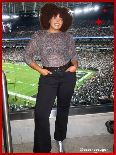 'short curly-haired woman stands in a stadium suite at night wearing a sheer silver sequin knit top over a black bra, black high-waisted pants with a black belt, and metallic silver ankle boots, with a packed football stadium field and crowd behind her, red star graphics in the top right corner and white username text @essencesugar_ in the bottom right corner'
