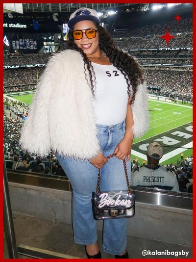 'Woman with long curly hair wears a white furry jacket over a white top with the number 52 on it, light-wash jeans, black heels, and a blue and white baseball cap with yellow-tinted glasses while holding a clear crossbody bag with a sparkly decorative front that reads Bebe in a stadium suite overlooking a football field; red stars sit in the top right corner of the frame. @kalanibagsby is written in the bottom right corner.'