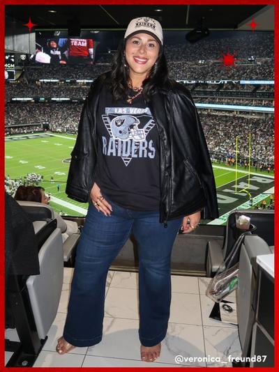 'Woman stands in a stadium suite wearing a black Las Vegas Raiders graphic tee under a black leather jacket, dark blue bootcut jeans, a tan Raiders baseball cap, hoop earrings, and open-toe sandals. She smiles at the camera with the football field and crowd visible behind her. Image has a red border with red starburst accents and the text @veronica_freund87 in the bottom right corner.'