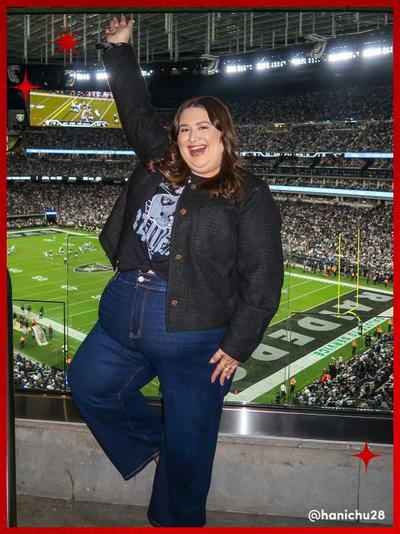 'Woman at a football stadium raising one arm and smiling, wearing a black Raiders graphic tee knotted at the waist under a textured black jacket with dark high-waisted wide-leg jeans, standing in front of a glass barrier overlooking the field with a crowd in the background and small red star graphics in the corners. @Hanichu26 is written in the bottom right corner.