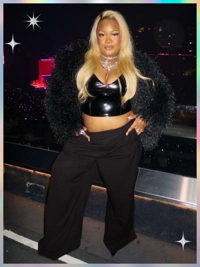 'A woman stands in front of a crowded concert arena wearing a black faux-fur jacket, a black vinyl crop top with a deep V neckline, and high-waisted wide-leg black pants; she has long straight blonde hair, layered silver necklaces, sculpted makeup with rosy cheeks and glossy lips, and her hands rest at her hips; white star-shaped sparkles appear in the top left and bottom right corners of the pastel-gradient frame'