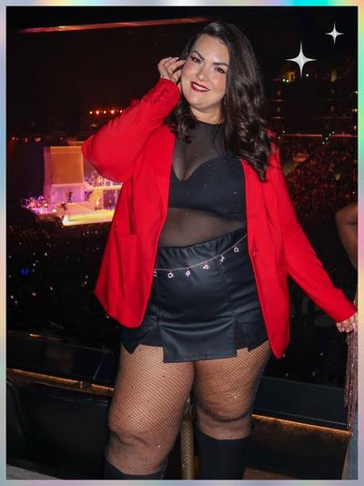 'A woman stands in front of a crowded concert arena wearing a bright red blazer over a sheer black mesh top with subtle sparkles, paired with a black faux-leather mini skirt featuring a small front slit and a silver chain belt; she wears black fishnet tights and black knee-high boots, and her dark wavy hair is styled loose; she poses with one hand touching her hair while white star-shaped sparkles appear in the upper right corner of the pastel-gradient frame'