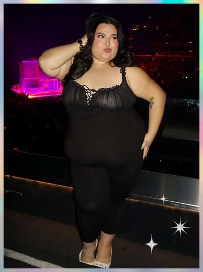 'A woman poses in front of a packed concert arena wearing a black sheer lace-trim babydoll-style top with a gathered bust and thin straps, paired with black fitted leggings; she has long wavy dark hair and wears cream-colored pointed-toe heels; she stands with one hand behind her head and the other at her side, with white star-shaped sparkles at the bottom right of the pastel-gradient frame'