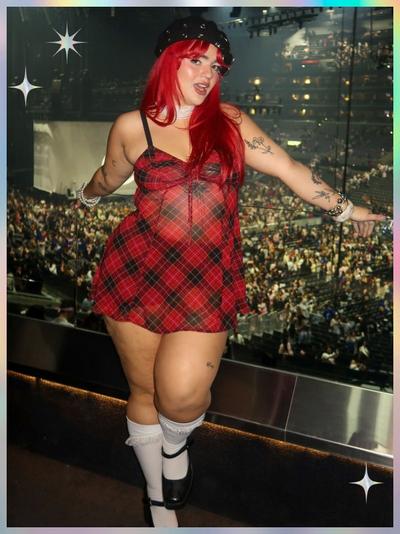 'A woman with long bright red hair poses in front of a large arena filled with people, standing against a glass barrier; she wears a sheer red and black plaid babydoll dress with black straps, a black beret decorated with silver studs, a white choker, white frilly knee-high socks, and black Mary Jane shoes; she has multiple tattoos on her arms including flowers and script, a bracelet on one wrist, and a nose ring, with white star-shaped sparkles in the corners of the image and a soft pastel border framing the scene'