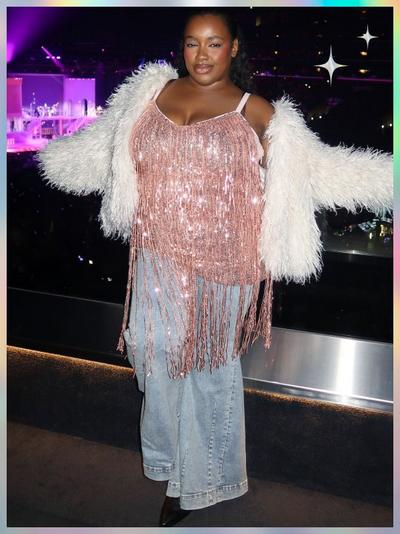 'A woman stands in front of a crowded arena, wearing a long pink sequined fringe tank layered over light-wash wide-leg jeans; she has a fluffy white jacket draped off her shoulders, dark pointed-toe shoes, and her hair is pulled back; star-shaped sparkles appear near her right shoulder with a soft pastel border framing the image'