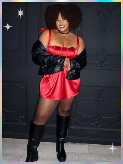 'A woman stands in front of a dark ornate wall wearing a short red satin dress with wide ribbon straps tied at the shoulders, a black choker necklace, red ball earrings, sheer black tights, and black knee-high boots; she has a large rounded afro hairstyle and a black leather jacket draped around her arms, with star-shaped sparkles in the upper left and lower right corners of the pastel-gradient frame'