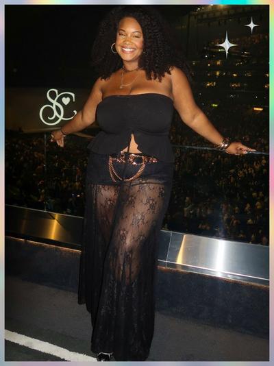 'A woman stands in an arena overlooking a large crowd, wearing a black strapless top with a center tie and a long sheer black lace skirt that reveals black shorts underneath; she has a rose-gold chain belt, hoop earrings, layered necklaces, bracelets, and voluminous curly hair, with white star-shaped sparkles in the top right corner of the pastel-gradient frame'