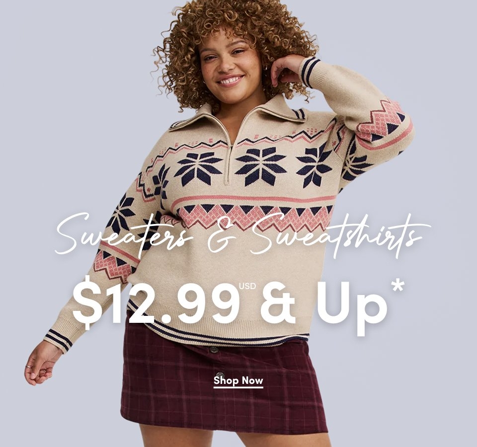 'A smiling woman with short tight curly light brown hair wears a beige quarter-zip sweater with navy and pink geometric snowflake and zigzag patterns across the chest and sleeves, ribbed cuffs with navy stripes, and a burgundy plaid mini skirt against a light gray background. White script text across the center reads Sweaters & Sweatshirts. Large white text across the lower center reads $12.99 USD & Up*. A white underlined Shop Now button appears below.'