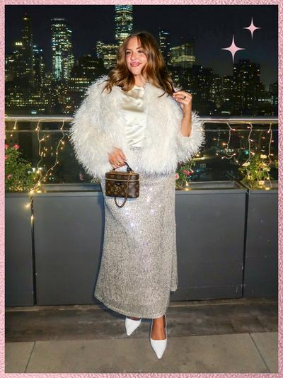 'A woman stands outdoors at night on a rooftop terrace in front of a city skyline, smiling and looking slightly to her left, wearing a white satin blouse with a high neckline, a cropped white shaggy faux-fur jacket, and a full-length silver sequined skirt, paired with pointed white heels, holding a small brown monogram handbag with a top handle, her light brown hair styled in loose waves, with string lights along a metal railing and flower boxes behind her, a concrete floor beneath her feet, and a pink textured border framing the image with pink sparkle star graphics in the upper right corner.'