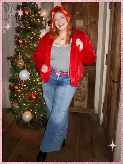 'A smiling woman stands on a wooden floor in front of a decorated Christmas tree with gold and white ornaments and warm lights, wearing a red faux-fur jacket over a gray scoop-neck top, blue jeans with a red ribbon belt, black shoes, and a red headband, with wood-paneled walls and a rustic wooden door behind her and pink sparkle star graphics in the corners of the image.'