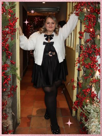 'A woman stands in a doorway framed with green garland and clusters of red berries, wearing a cream textured cardigan over a black dress with a large black bow at the neckline and a second bow at the waist, a black belt with a gold buckle, black tights, and black flats, with shoulder-length light brown hair styled in loose waves, both arms raised to hold the doorframe, warm string lights and holiday decor visible inside behind her, terracotta tile flooring underfoot, and pink sparkle star graphics near the upper left and lower right edges of the image.'