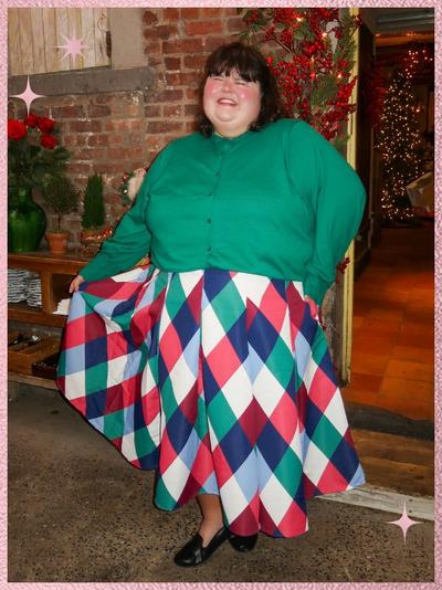 'A woman stands indoors smiling with eyes closed, wearing a forest green long-sleeve button-front blouse tucked into a full A-line midi skirt with a large diamond harlequin pattern in red, pink, teal, navy, and white, holding the skirt out with one hand, wearing black flats, with straight dark hair and blunt bangs, standing on a speckled concrete floor beside a wooden cabinet and exposed brick wall, a doorway behind her decorated with green garland and red berries leading to a warmly lit room with holiday lights, and pink sparkle star graphics near the upper left and lower right edges of the image.'
