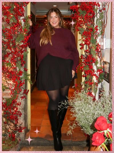 'A woman stands smiling in a doorway framed with green garland and red berries, wearing a deep burgundy knit sweater with relaxed sleeves tucked into a black flared mini skirt, sheer black tights, and black knee-high boots, with long brown hair styled loose and parted at the center, one hand resting on the doorframe, standing on terracotta tile flooring with warm holiday lights visible inside behind her, frosted greenery and red flowers arranged at the lower right, a pink textured border framing the image, and small pink sparkle star graphics near the lower left corner.'