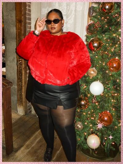 'A woman stands indoors beside a decorated Christmas tree with warm white lights and copper, gold, and white round ornaments, wearing a bright red faux fur cropped jacket, a black faux leather mini skirt, sheer black tights, and black slip-on shoes with small metallic studs, accessorized with black cat-eye sunglasses, gold hoop earrings, and a bracelet, her hair pulled back, one hand raised to adjust her sunglasses, standing on a wooden floor near a brick wall and doorway with lace curtains, a pink textured border framing the image with small pink sparkle star graphics near the lower right.'
