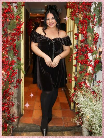 'A woman stands centered in a doorway decorated with red berry garlands and evergreen branches, smiling at the camera, wearing a black off-the-shoulder velvet mini dress with a small bow tie detail at the neckline, sheer black tights, and black pointed-toe heels, her dark hair styled half up with volume at the crown and loose curls framing her face, hands clasped at her waist, standing on terracotta tile flooring with warm string lights and holiday decor visible inside the room behind her, framed by a pink textured border with small pink sparkle star graphics near the lower left and mid right edges.'
