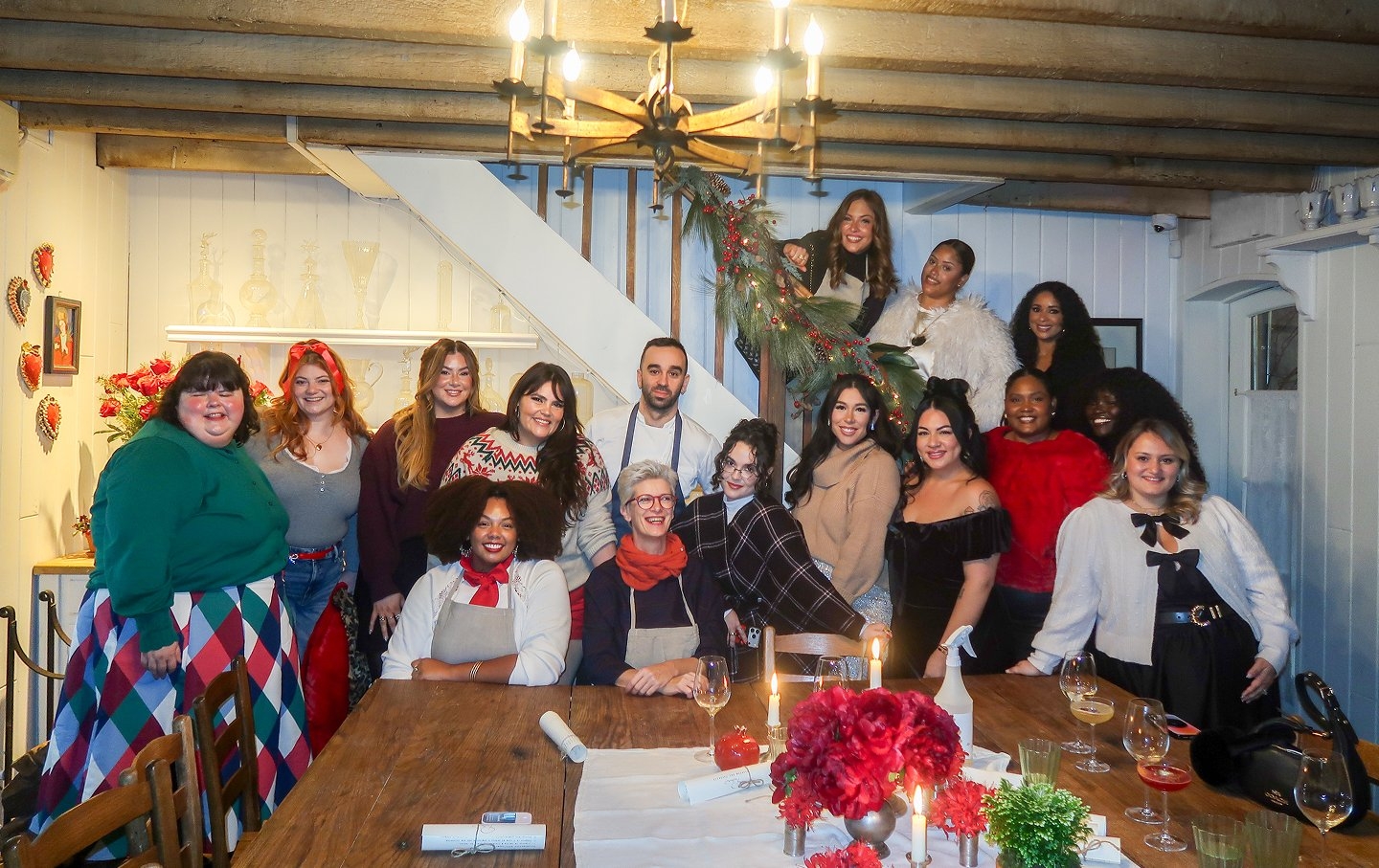 'Large group of people posing together in a cozy rustic dining room decorated for the holidays, standing and sitting around a long wooden table with lit candles, wine glasses, rolled place cards, and red floral centerpieces, exposed wooden ceiling beams and a chandelier overhead, white walls with shelves of glassware, a staircase decorated with greenery and red accents behind them, and everyone wearing festive winter outfits in reds, greens, neutrals, and patterns, smiling toward the camera.'