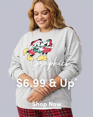 A medium-shot photograph of a light-skinned model with wavy blonde hair smiling and looking down. She is wearing a grey crewneck sweatshirt featuring a graphic of Mickey and Minnie Mouse in holiday attire. She is also wearing red and black plaid pajama pants. Overlaid on the image is the word 'Graphics' in a white cursive script, followed by '$6.99 USD & Up*' in a large, bold white sans-serif font. A small white underlined 'Shop Now' link is centered at the bottom.