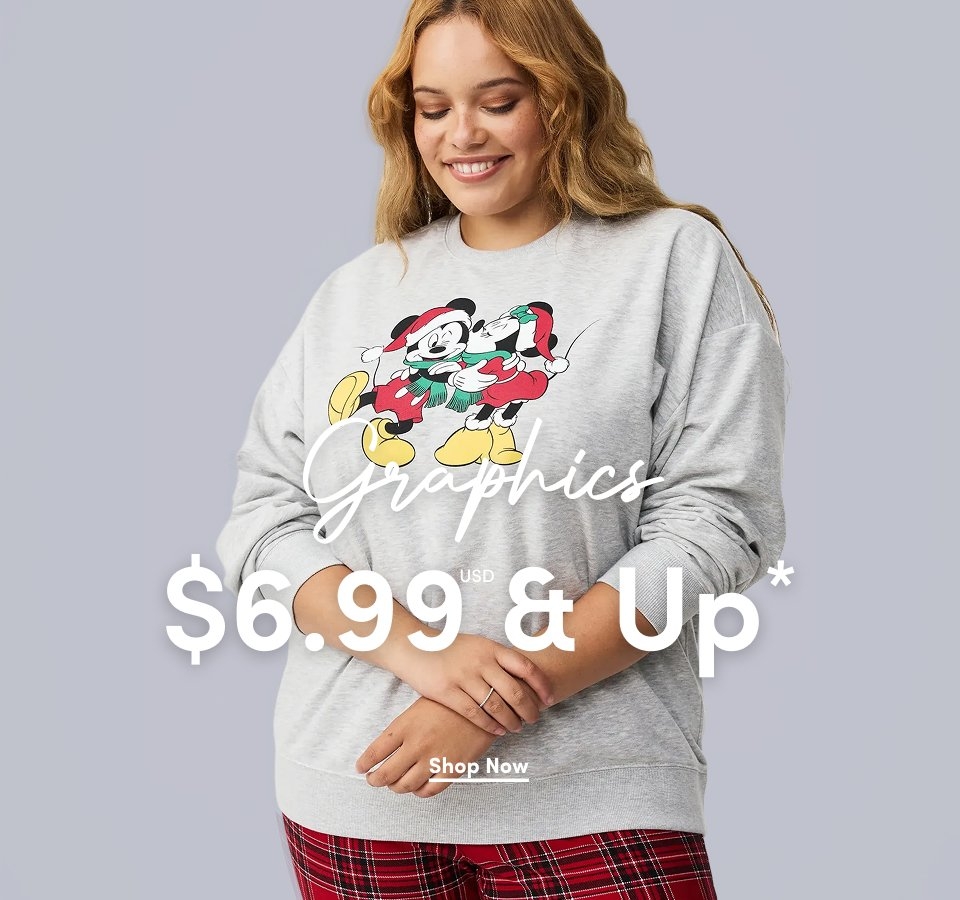 A medium-shot photograph of a light-skinned model with wavy blonde hair smiling and looking down. She is wearing a grey crewneck sweatshirt featuring a graphic of Mickey and Minnie Mouse in holiday attire. She is also wearing red and black plaid pajama pants. Overlaid on the image is the word 'Graphics' in a white cursive script, followed by '$6.99 USD & Up*' in a large, bold white sans-serif font. A small white underlined 'Shop Now' link is centered at the bottom.