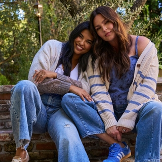 A lifestyle photograph of two plus-size models sitting closely together on brick steps in an outdoor garden setting. The model on the left smiles while wearing a white shirt, a speckled brown and white knit cardigan, and light-wash jeans. The model on the right leans her head against the other, wearing a blue floral camisole under a cream cardigan with blue horizontal stripes on the sleeves. A vintage lamp post and green foliage are visible in the soft-focus background.