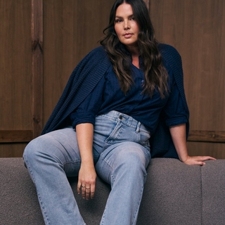 A plus-size model with long, wavy brown hair sits on a grey textured surface against a dark wood-paneled wall. She is wearing a navy blue button-down shirt tucked into light-wash jeans, with a chunky navy blue knit cardigan draped over her shoulders. She looks directly at the camera with a neutral expression.