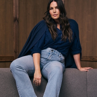 A plus-size model with long, wavy brown hair sits on a grey textured surface against a dark wood-paneled wall. She is wearing a navy blue button-down shirt tucked into light-wash jeans, with a chunky navy blue knit cardigan draped over her shoulders. She looks directly at the camera with a neutral expression.