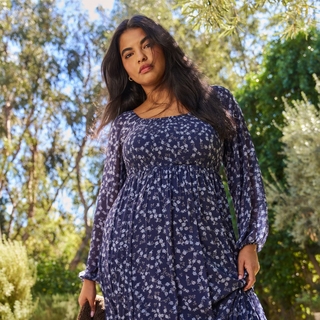 A low-angle lifestyle photograph of a plus-size model with long, dark hair standing outdoors against a backdrop of sunlit trees. She is wearing a navy blue maxi dress with a small white floral print, featuring a smocked bodice and long sheer sleeves. She looks down toward the camera with a soft expression.