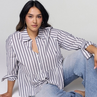 A plus-size model with long, dark hair sits on a white floor against a white background. She is wearing a blue and white vertical-striped button-down shirt with a ruffled peplum hem and sleeves rolled up to the elbows, paired with light-wash jeans. She is leaning back slightly with one hand resting on her knee, looking directly at the camera.