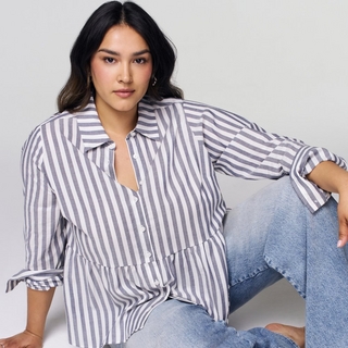 A plus-size model with long, dark hair sits on a white floor against a white background. She is wearing a blue and white vertical-striped button-down shirt with a ruffled peplum hem and sleeves rolled up to the elbows, paired with light-wash jeans. She is leaning back slightly with one hand resting on her knee, looking directly at the camera.