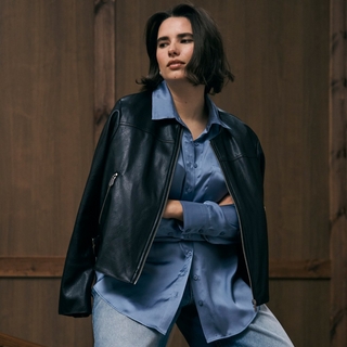 A plus-size model with short dark hair stands against a dark wood-paneled wall. She is wearing a light blue satin button-down shirt tucked into light-wash jeans, with a black faux-leather motorcycle jacket draped over her shoulders. She has her arms crossed and is looking off to the side with a calm expression.